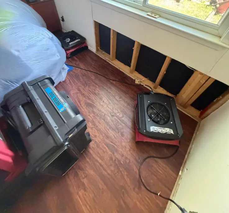 A room undergoing water damage restoration, with a section of the wall below a window removed to expose insulation. Two air movers, a HEPA 700 air scrubber and a Phoenix Airmax, are in use on the brown wood-look flooring.